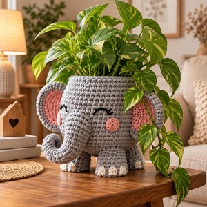 Puede incluir: Una maceta de elefante tejida a ganchillo en color gris con orejas grandes de centro rosa, trompa curvada y ojos negros bordados. El elefante tiene detalles blancos en los pies y mejillas rosas, conteniendo una planta pothos verde y amarilla.