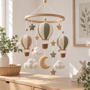 Peut inclure: Un mobile de chambre d'enfant composé d'un cadre circulaire en bois avec des éléments en crochet suspendus, incluant trois montgolfières vert sauge, crème et marron, des nuages crème, un croissant de lune jaune et des étoiles vert et beige.