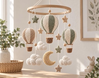 Op de afbeelding: Een babymobiel met een rond houten frame en hangende gehaakte elementen, waaronder drie heteluchtballonnen in saliegroen, crème en bruin, diverse crèmekleurige wolken, een gele halve maan en verschillende kleine sterren in groen en beige.