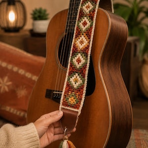 Op de afbeelding: Een bruine akoestische gitaar met een gehaakte gitaarband met een ruitpatroon in roest, olijfgroen en crème. De band is bevestigd aan een bruin lederen uiteinde met een veelkleurige kwast in groen, crème en roest die eraan hangt.