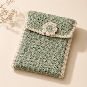 May include: A sage green rectangular crochet pouch featuring a fold-over flap with a cream-colored crocheted flower embellishment at the center. The edges of the pouch and the flap are finished with a cream-colored border in a simple stitch.