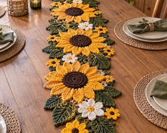 Op de afbeelding: Een lange, rechthoekige gehaakte tafelloper met een herhalend patroon van grote gele zonnebloemen met donkerbruine harten, afgewisseld met kleinere witte madeliefachtige bloemen en gestructureerde donkergroene bladeren in een bloemendesign.