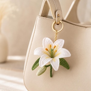 Può includere: Un portachiavi a forma di fiore di giglio bianco realizzato in filo all'uncinetto, con stami gialli e foglie verdi, fissato a un anello e una catena in metallo dorato, appeso a una borsa color crema con superficie liscia e strutturata.
