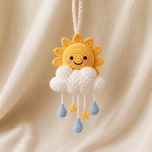 Può includere: Un ornamento da appendere all'uncinetto raffigurante un sole giallo con un viso sorridente sopra una nuvola bianca. Tre fili bianchi pendono dalla nuvola, ciascuno terminante con una goccia di pioggia azzurra o una piccola stella gialla, con un cappio.