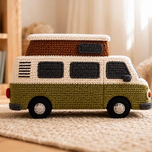 May include: A crocheted camper van toy featuring an olive green lower body, a cream-colored upper section, and a brown pop-up roof. The vehicle includes dark gray windows, black wheels with light gray centers, a red taillight, and a small orange headlight.