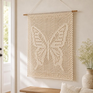 Peut inclure: Une tenture murale rectangulaire en crochet de couleur crème représentant un grand motif de papillon au centre. La pièce est suspendue à une tige en bois horizontale par une ficelle blanche et présente un motif en grille ajouré complexe.