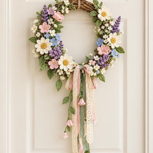 May include: A circular wreath constructed from brown woven twigs, adorned with crocheted flowers in shades of white, pink, blue, and purple, along with green leaves. A cluster of hanging ribbons in cream, pink, and sage green drapes from the bottom.