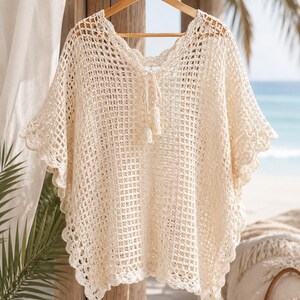 May include: An off-white crochet cover-up featuring an open grid pattern, a V-neckline with a drawstring tie finished with two tassels, and scalloped edges along the hem and sleeves. The garment is displayed on a wooden hanger.