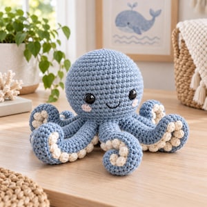 May include: A blue crocheted octopus plush toy with a round head, black plastic eyes, a stitched smile, and pink blush accents. The underside of each of the eight curled tentacles features small, cream-colored crocheted circular suction cups.