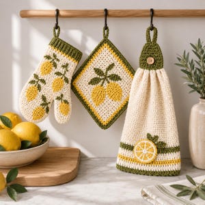 Pode incluir: Um conjunto de cozinha em crochê composto por uma luva de forno, um descanso de panela quadrado e uma toalha de pendurar. Cada peça é creme com acabamento verde e motivos de limões amarelos. A toalha inclui um botão de madeira central.