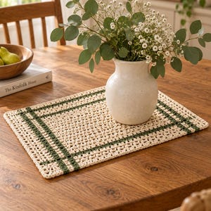 Op de afbeelding: Een rechthoekige gehaakte placemat met een cr&egrave;mekleurig opengewerkt midden en een donkergroene rand met een dubbel lijnpatroon. De placemat is gemaakt met een getextureerde garenfinish en vertoont een consistent, herhalend geometrisch steekpatroon.