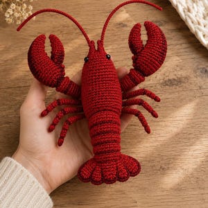 May include: A deep red crochet lobster toy featuring a segmented body, two large front claws, multiple thin legs, and two long antennae. The lobster has small black bead eyes and is crafted with a detailed, textured yarn stitch pattern throughout.
