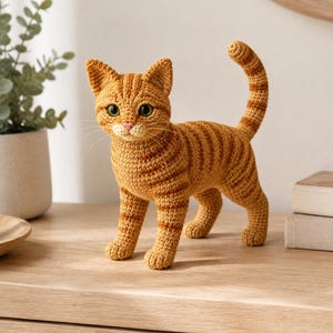 May include: A crocheted orange tabby cat figurine with dark orange stripes, green eyes, and a pink nose. The cat is standing with its tail held high, featuring detailed yarn stitching throughout its body, pointed ears, and long white whiskers.