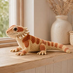 Può includere: Un peluche a forma di drago barbuto realizzato all'uncinetto, caratterizzato da un corpo color crema con macchie arancioni-marroni, una barba spinosa sulla gola e occhi marrone scuro. La lucertola è lavorata con punti dettagliati.