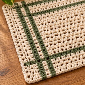 Op de afbeelding: Een rechthoekige gehaakte mat met een gebroken witte basis en een rasterachtig patroon van donker bosgroen garen. Het textiel is gemaakt met een opengewerkt steekontwerp, wat zorgt voor een gestructureerd oppervlak met zichtbare vierkante openingen.