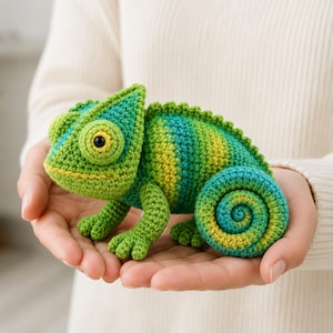 Puede incluir: Un juguete de camaleón tejido a ganchillo con una mezcla vibrante de hilo verde y amarillo. El peluche tiene una superficie texturizada, un ojo prominente con pupila negra e iris amarillo, y una cola enroscada en forma de espiral.