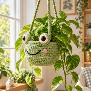 May include: A light green crochet hanging planter shaped like a frog with large black and white eyes, pink cheeks, and a black stitched smile. The planter features two long braided straps for suspension and holds a trailing green pothos plant.