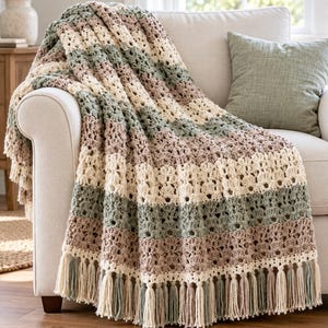 May include: A crocheted throw blanket featuring horizontal stripes in alternating shades of cream, sage green, and dusty rose. The blanket has a textured, open-stitch pattern and is finished with long, matching yarn tassels along the bottom edge.