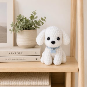 May include: A white crocheted stuffed dog toy with black plastic eyes and a black nose. The dog wears a light blue collar with a small bone-shaped tag. The plush toy is positioned on a wooden shelf next to a potted green plant and two books.