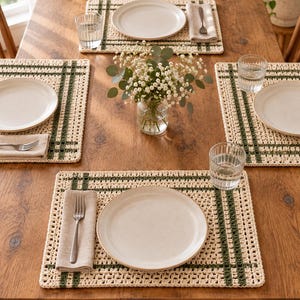 Op de afbeelding: Een set rechthoekige gehaakte placemats met een cr&egrave;mekleurige basis en donkergroene strepen in ruitpatroon. Elke placemat is op een houten tafel gerangschikt met een wit keramisch bord, een beige linnen servet, een zilveren vork en een glas.