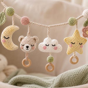 May include: A crochet garland featuring hanging figures including a yellow crescent moon, a beige bear face, a white cloud, and a yellow star, all with embroidered sleepy faces and pink cheeks, interspersed with small green spheres and wooden rings.