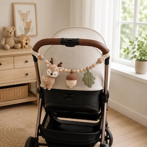 Könnte beinhalten: Ein Kinderwagenspielzeug mit einer Schnur aus hellen Holzperlen und drei gehäkelten Figuren: ein rehbraunes und weißes Reh mit braunem Geweih, eine braun-cremefarbene Eichel und ein grünes Eichenblatt, befestigt am braunen Ledergriff des Wagens.