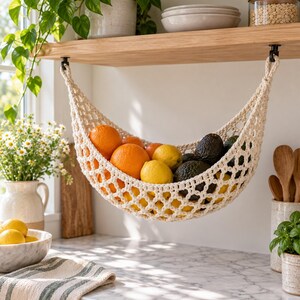Puede incluir: Una hamaca para frutas de macramé color crema suspendida de un estante de madera, llena de una variedad de productos frescos que incluyen naranjas brillantes, limones amarillos y aguacates verde oscuro, como solución de almacenamiento.