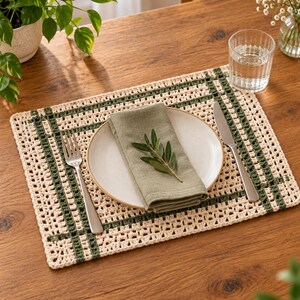 Op de afbeelding: Een rechthoekige gehaakte placemat met een cr&egrave;mekleurig opengewerkt patroon en donkergroene rasterlijnen. In het midden staat een rond keramieken bord, met daarop een gevouwen saliegroene linnen servet, een klein olijftakje, een zilveren vork en een mes.