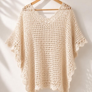 May include: A cream-colored crochet poncho featuring an open grid pattern, a V-neckline, and scalloped edges along the hem and sleeves. The garment is constructed with a lightweight, airy yarn and hangs from a wooden hanger, displaying a loose fit.