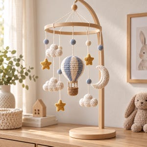 Peut inclure: Un mobile de chambre d'enfant avec un support en bois clair et des éléments en crochet suspendus, incluant une montgolfière centrale, des nuages blancs, des étoiles jaunes, un croissant de lune et de petites sphères bleues sur des fils.