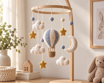 Op de afbeelding: Een babykamer mobiel met een standaard van licht hout en hangende gehaakte elementen, waaronder een centrale heteluchtballon, witte wolken, gele sterren, een halve maan en kleine blauwe bolletjes aan witte koordjes aan een houten ring.