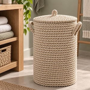 May include: A cylindrical laundry basket made of thick, cream-coloured woven rope. The basket features a matching lid with a small central handle and two sturdy side handles. The texture is uniform and dense, created by a continuous crochet-style stitch.