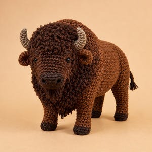 Puede incluir: Un bisonte de ganchillo hecho a mano, peluche con una melena texturizada de color marrón oscuro, un cuerpo de color marrón claro y dos pequeños cuernos grises. El peluche tiene ojos de cuentas negras, una cola corta y cuatro patas con pezuñas.