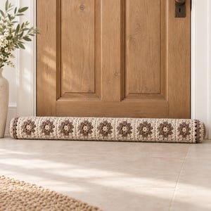 May include: A long, cylindrical door draft stopper featuring a crocheted pattern of repeating beige and brown floral motifs. The ends are finished with solid brown circular caps, and the overall design is consistent across the entire length.