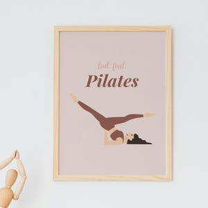 May include: A framed wall art print featuring a light mauve background with the text but first Pilates in dark brown letters above an illustration of a figure performing a Pilates pose in brown athletic wear, alongside a wooden artist mannequin.