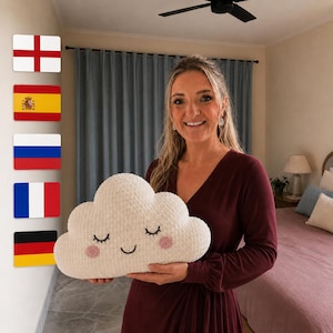 Puede incluir: Un cojín de peluche en forma de nube de color crema con una cara bordada que muestra ojos cerrados, una pequeña sonrisa y dos mejillas circulares de color rosa. El cojín tiene una superficie texturizada y se sostiene frente a una persona.