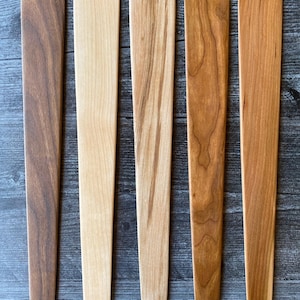 May include: Five flat wooden kitchen spatulas arranged vertically in a row. Each spatula features a distinct wood grain pattern and color, ranging from a dark brown walnut tone to lighter shades of maple and cherry wood with smooth, rounded edges.