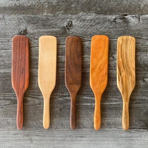 May include: Five wooden spatulas arranged in a row, each featuring a distinct wood grain pattern and color, ranging from light tan to deep reddish-brown and dark brown, with flat, rectangular heads and smooth, rounded handles for kitchen use.