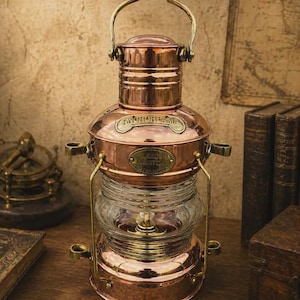 May include: A polished copper ship lantern featuring a clear glass centre section, a brass handle, and two oval metal plaques. The top plaque reads ANCHOR LAMP, and the lower plaque reads LEEDS BURTON. The lantern has a cylindrical body with brass accents.