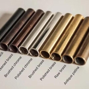 May include: A collection of nine cylindrical metal tubes arranged in a row, each featuring a distinct finish. The labels include Matte black, Antique black, Blackened brass, Brushed chrome, Polished chrome, Brushed brass, Polished brass, Raw brass, and Antique patina.