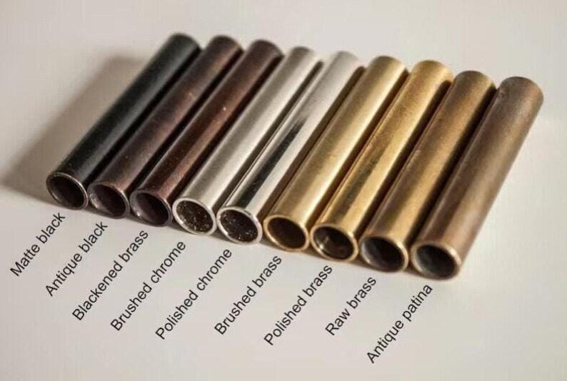 May include: A collection of nine cylindrical metal tubes arranged in a row, each featuring a distinct finish. The labels include Matte black, Antique black, Blackened brass, Brushed chrome, Polished chrome, Brushed brass, Polished brass, Raw brass, and Antique patina.