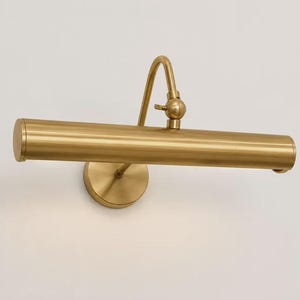 May include: A wall-mounted picture light with a brushed brass finish. The fixture features a horizontal cylindrical lamp head attached to a curved arm with an adjustable swivel joint, connected to a circular base plate mounted to the wall.
