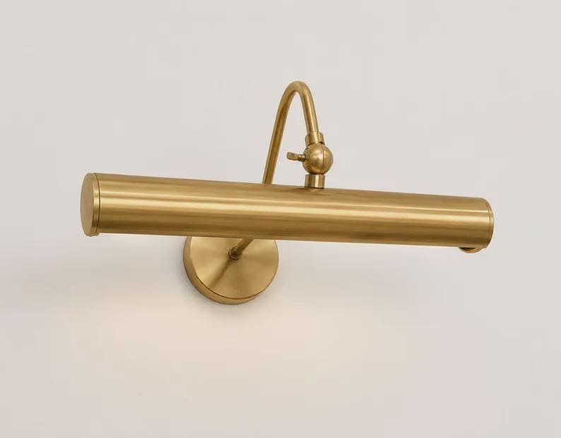 May include: A wall-mounted picture light with a brushed brass finish. The fixture features a horizontal cylindrical lamp head attached to a curved arm with an adjustable swivel joint, connected to a circular base plate mounted to the wall.
