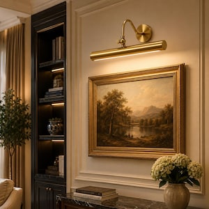 May include: A brass picture light fixture with a cylindrical head and an adjustable arm mounted above a rectangular landscape painting in a gold frame. The light emits a warm glow downward, illuminating the artwork on the wall.