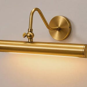 May include: A wall-mounted picture light with a brushed brass finish. The fixture features a cylindrical horizontal lamp head attached to a curved adjustable arm and a circular wall plate. A warm light emits from the bottom of the tube.