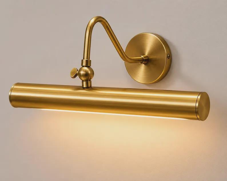 May include: A wall-mounted picture light with a brushed brass finish. The fixture features a cylindrical horizontal lamp head attached to a curved adjustable arm and a circular wall plate. A warm light emits from the bottom of the tube.