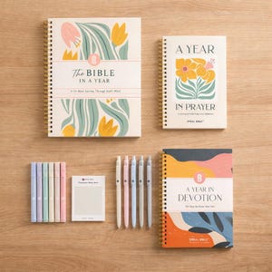 May include: An assortment of stationery items, including spiral-bound journals and pens. The journals feature floral designs and titles like "The Bible in a Year" and "A Year in Devotion." Pastel highlighters and sticky notes are also included.