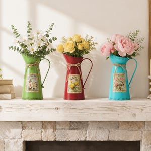 Può includere: Tre brocche di metallo colorate con fiori. Le brocche sono verdi, rosse e turchesi, ognuna con un'etichetta floreale e un fiocco di spago. La brocca verde contiene margherite bianche, la brocca rossa ha rose gialle e la brocca turchese contiene peonie rosa.