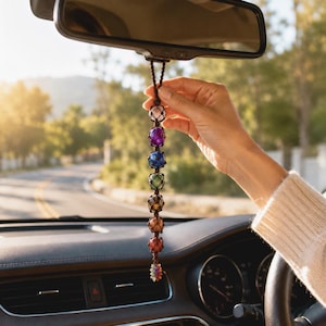 Puede incluir: Un adorno para coche con una cadena de cuentas de piedras preciosas de colores en tonos rosa, morado, azul, verde, marrón, naranja y rojo. El adorno está suspendido de un cordón marrón y cuelga del espejo retrovisor de un coche.