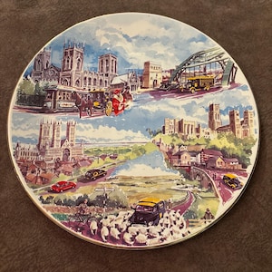 May include: A round decorative ceramic plate featuring a colorful illustration of a cityscape with historic stone buildings, a large arched bridge, vintage automobiles, a horse-drawn carriage, and a flock of sheep grazing in the foreground.